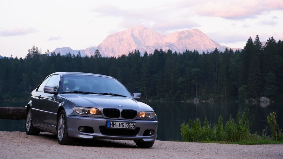Γκρι BMW 3 Series E46 παρκαρισμένη δίπλα σε λίμνη με βουνά.