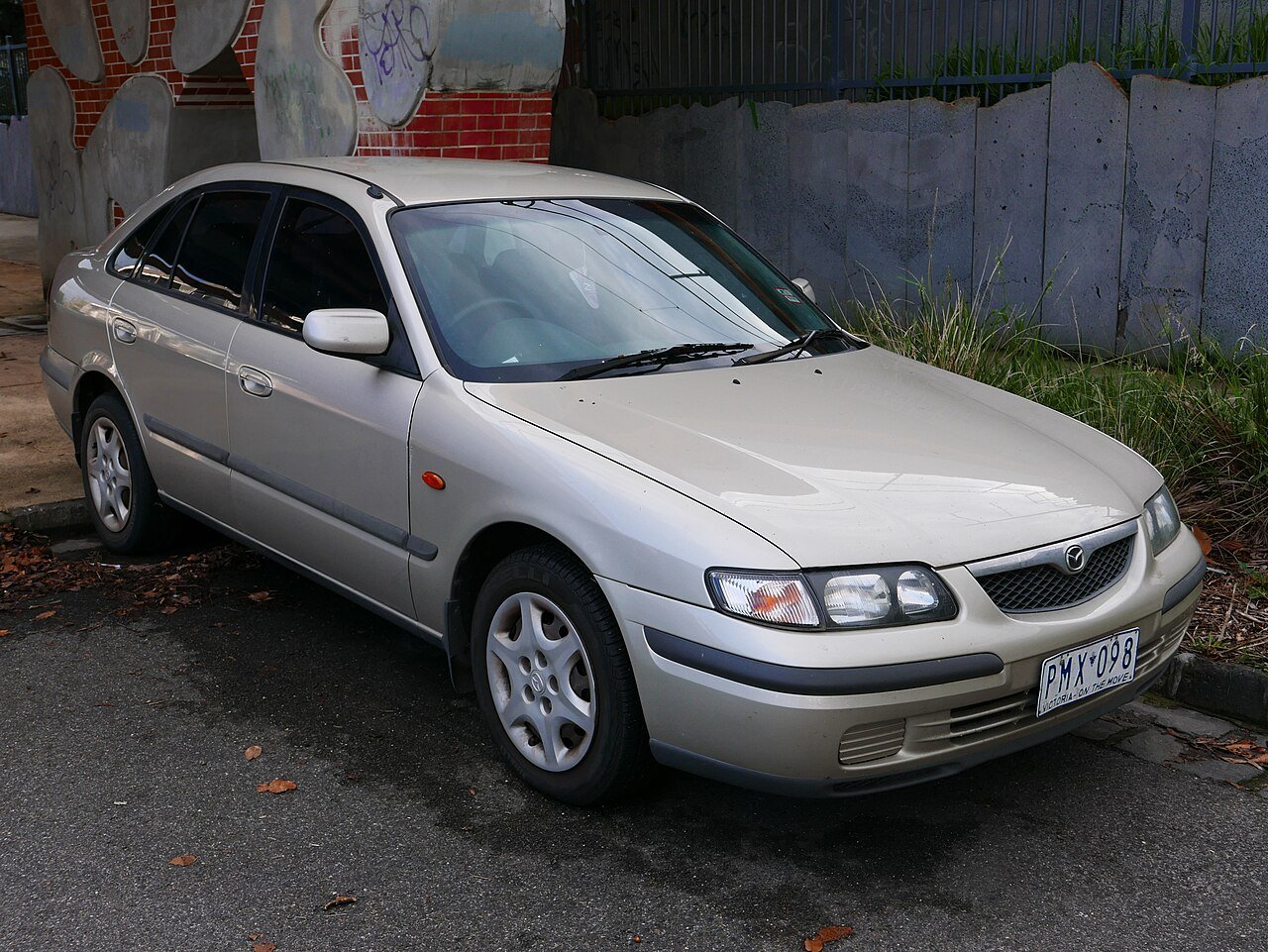 Χρυσό Mazda 626 σταθμευμένο στο δρόμο, με φόντο έναν τοίχο.